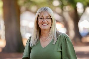 A woman in a green v-neck sweater smiles warmly while standing outdoors with trees in the background.