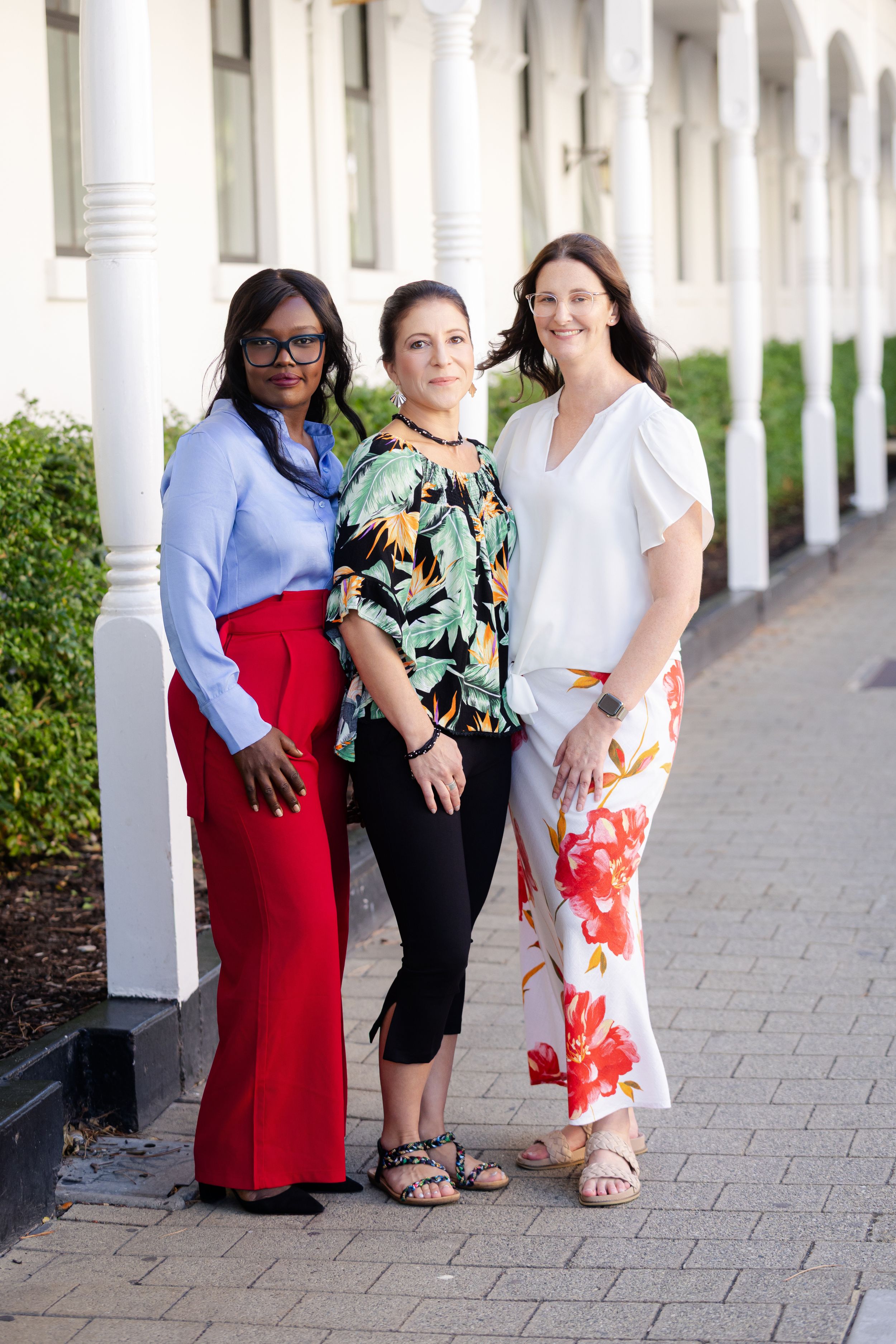 Three colleagues in colorful professional attire pose together against a white colonnade walkway with hedges.