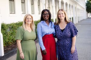 Three coworkers in business casual attire smile while standing outside an office building with white columns.