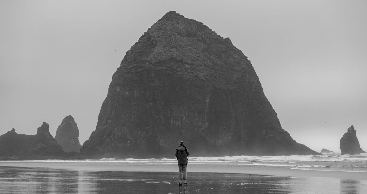 Black and white photo of a lone figure walking along the beach at Haystack Rock, Oregon. Moody skies & wet sand create a cinematic scene.