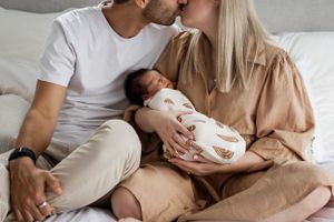 A tender family moment together on a cozy bed as they gaze at their sleeping newborn wrapped in patterned swaddle.