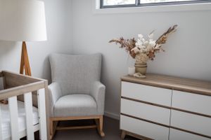 Modern nursery corner with gray armchair, white crib, and wooden dresser decorated with dried flowers.