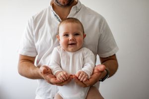 A baby in white clothing smiling while being held against a white background.