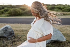 Expectant mother in white dress poses during sunset with hair blowing in the wind.