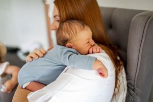 Parent hugging sleeping baby in light blue onesie on grey armchair.