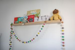Children's books and a plush toy displayed on a floating shelf with colorful pom pom garland.