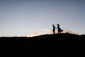A silhouetted couple stands holding hands on a hilltop against a sunset sky.