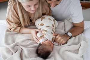 Parents sharing an intimate moment looking down at their swaddled newborn on beige blanket.