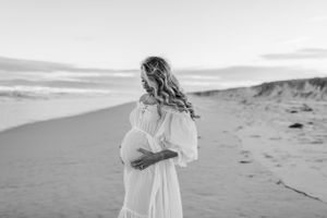 A woman in a flowing white dress poses on a beach dune at dusk.