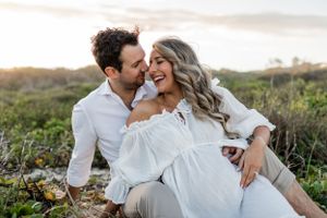 A romantic couple laughs together while sitting in a grassy field during golden hour.