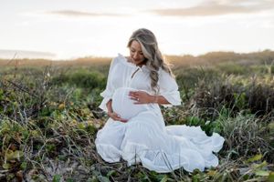 Woman in white dress sitting in field of wildflowers during sunset maternity shoot.