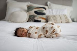 Sleeping infant wearing rainbow patterned swaddle rests peacefully on white bed.