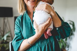 Woman in teal dress holding and gazing lovingly at newborn baby indoors.