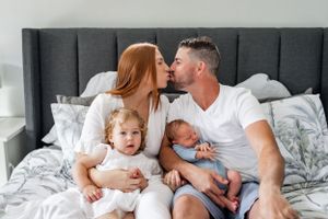 Intimate family moment with toddler and newborn on bed in white bedroom.