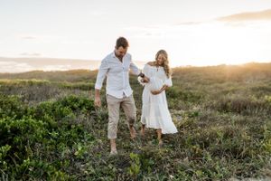 A couple in white outfits walks hand in hand through a scenic meadow at sunset.