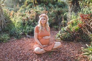 A maternity photo of an expectant mother in an orange dress sitting on a garden path surrounded by lush plants.