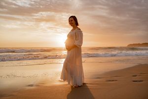 A pregnant figure in a flowing white dress stands on a beach at golden sunset while waves crash on the shore.