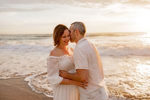 A couple embraces on a beach at sunset, with gentle waves lapping at the shore and warm golden light.