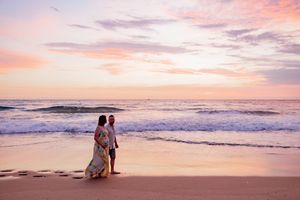 Two people stroll along a sandy beach at sunset with pink and orange skies reflecting on the ocean waves.