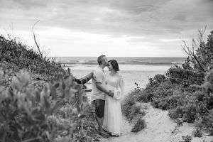 Black and white beach wedding photo showing two people embracing on coastal path with ocean view in background.