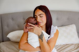 Mother in white top holds newborn baby lovingly on bed with gray headboard.