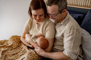 A couple sits on a couch with a newborn baby while covered in a floral patterned blanket.