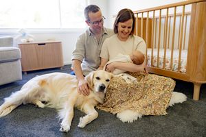 A couple sits with their newborn baby in a nursery while a white dog rests beside them on the floor.