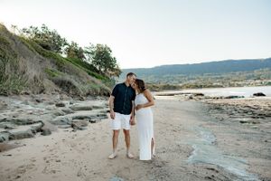 Expecting couple walking together on a scenic rocky beach with cliffs and mountains in background.