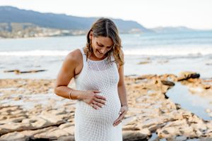 Pregnant woman in white crochet dress poses on rocky coastline with ocean and mountains behind.