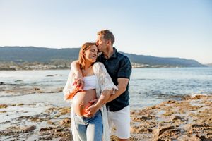 Romantic couple's maternity photo on rocky beach during golden hour with coastal backdrop.