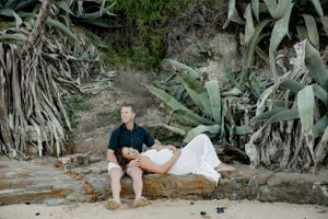 A couple rests on rocky terrain surrounded by large coastal plants.