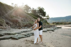 A couple embraces on a rocky beach with hills and cliffs in the background.