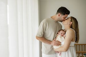 A tender family moment as loving parents embrace while holding their sleeping newborn baby near a bright window.