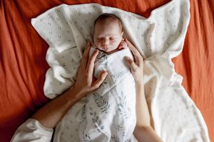 Newborn baby wearing white clothing lies on an orange background with white blanket.