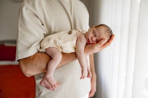 A sleeping newborn baby in cream-colored onesie rests peacefully on someone's forearm by a window with white curtains.