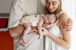 A sleeping baby wearing a cream-colored onesie rests peacefully against their parent's chest at home.