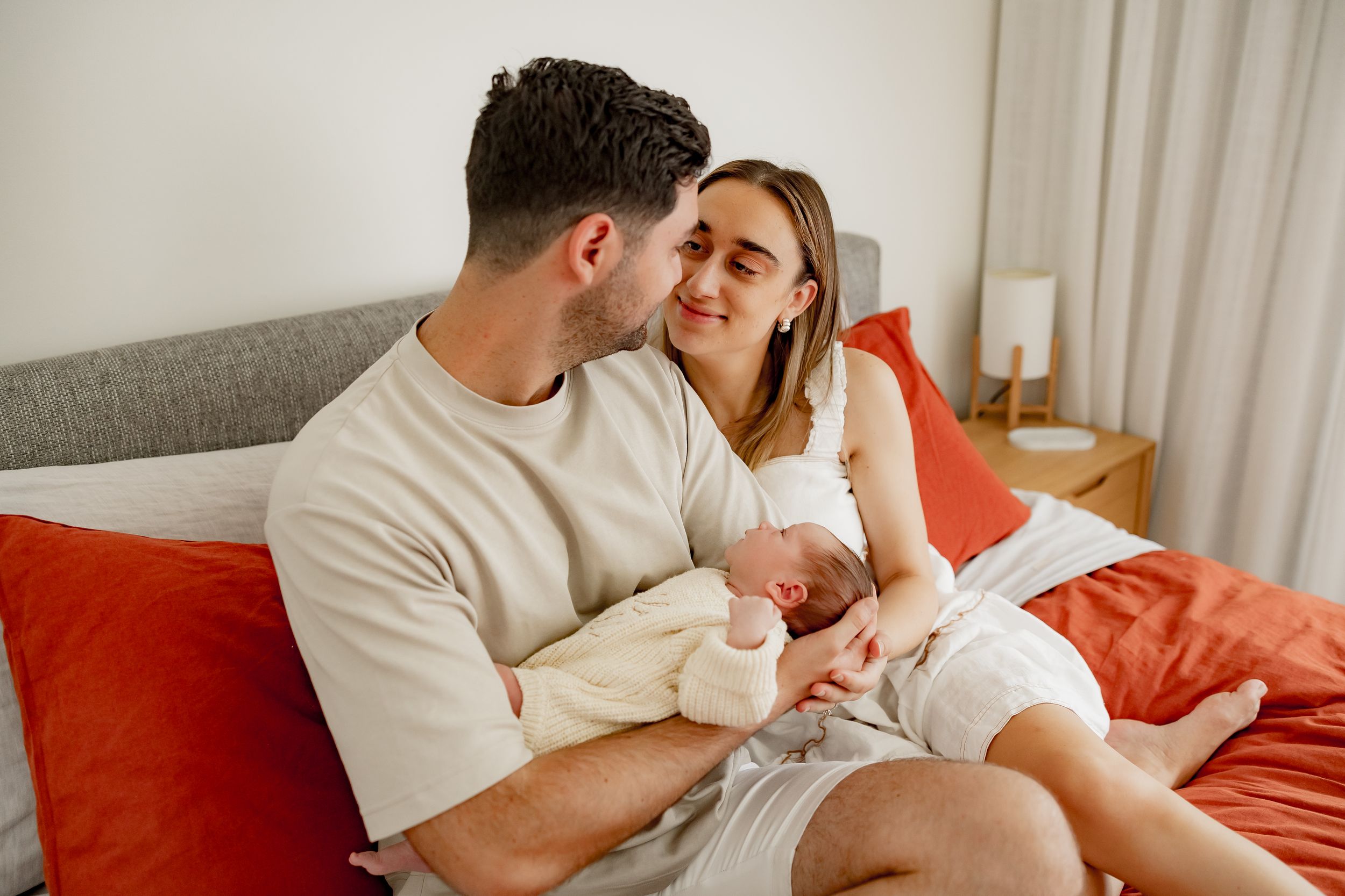 Couple in white lounging on gray couch with red throw pillows in bright bedroom.
