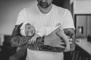 Black and white photo of newborn baby being cradled in tattooed arms.