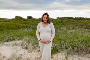 Expectant mother in white dress poses in natural coastal landscape.