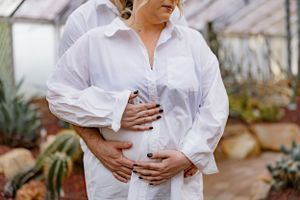 Elegant white maternity photo features billowing sleeves and gentle hands cradling a baby bump in a greenhouse setting.
