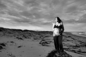 Black and white maternity photo of pregnant woman standing on sandy beach under dramatic sky.