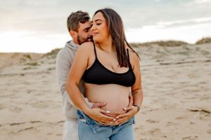 Romantic maternity beach photo with couple embracing at sunset on sandy dunes.