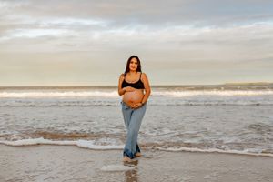 Pregnant woman in denim and black top poses by ocean waves during beach photoshoot.