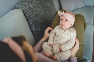 A baby wearing a cream sweater and bow headband sits in a cozy armchair.