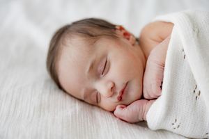 A sleeping newborn baby wrapped in a soft white blanket rests peacefully on a light-colored background.