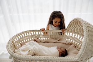 Child looking down at baby sibling resting in white wicker bassinet near window.