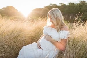 Pregnant woman in white dress sitting in golden sunlit field at sunset.