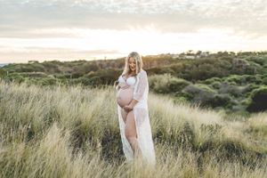 Expecting mother in white dress standing among tall grass during golden hour.