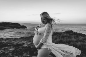 A maternity photoshoot at a rocky coastal shoreline in black and white with flowing lace dress and windswept hair.