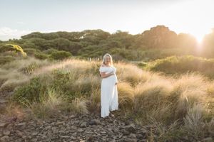 Ethereal maternity photo in long white dress amid golden sunlit coastal grasses.